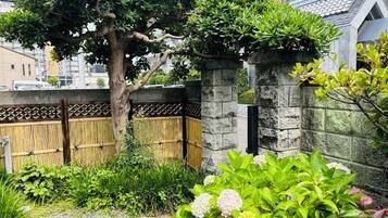 Um jardim japonês tradicional com parede de pedra, cerca de bambu e diversas plantas.