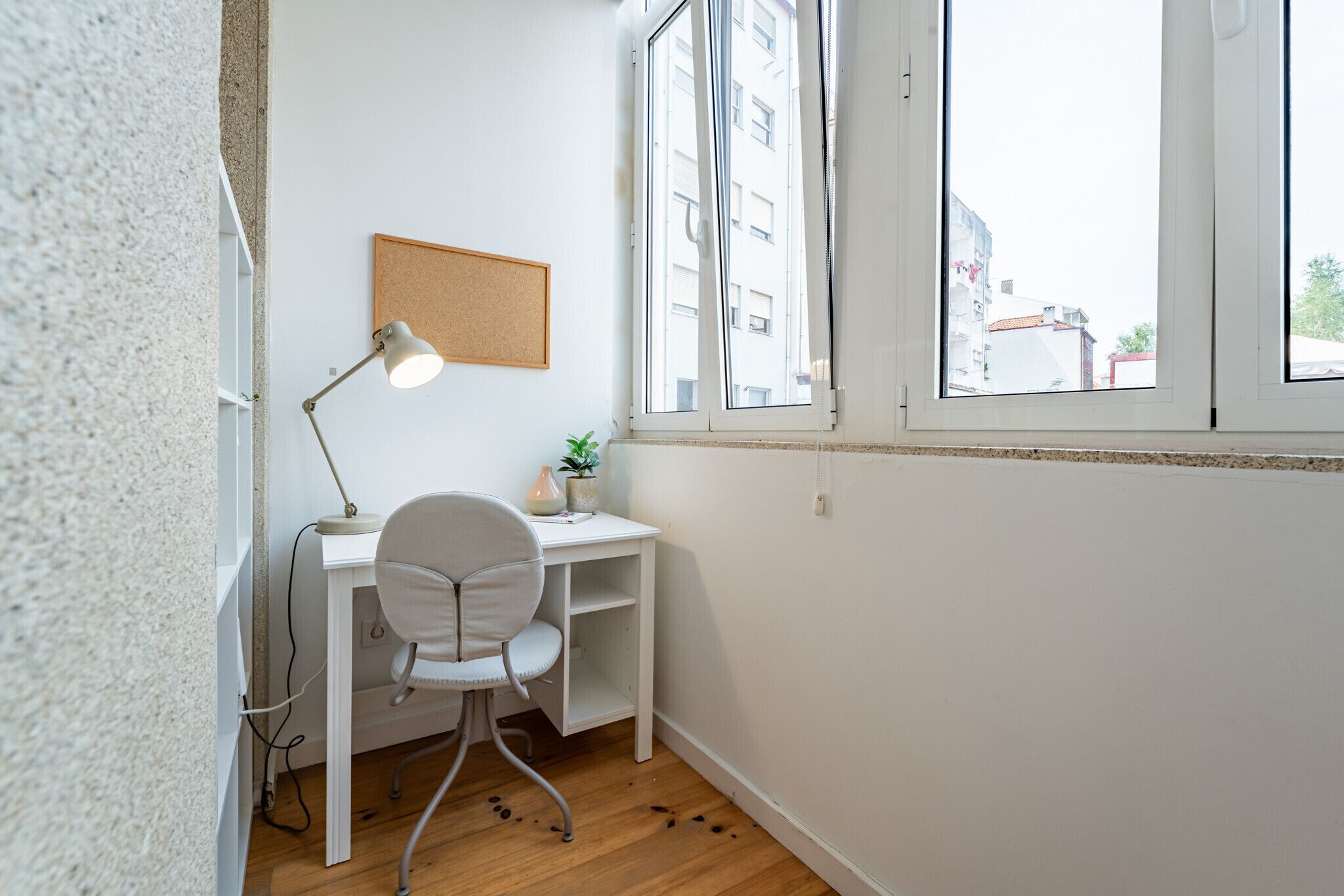 Een kleine, nette kamer met een bureau, stoel en een raam met uitzicht op gebouwen.