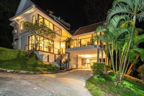 A modern two-story house with a well-lit entrance, a visible staircase, and a lush garden with palm trees. - patong hill 3 villa (Phuket)