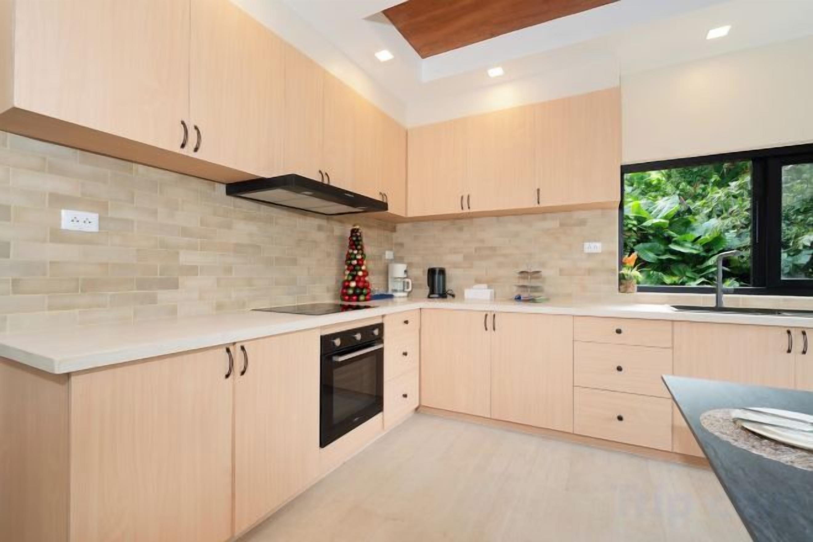 A modern kitchen with light wood cabinets, a built-in oven, and a window providing a view of greenery.