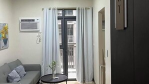 A living room with a grey sofa, a small round table with a plant, a window with curtains, an air conditioning unit, and a door with a sign. - Near MOA beside Okada Pet Friendly 1BR 3 pax WIFI 8T (Paranaque)