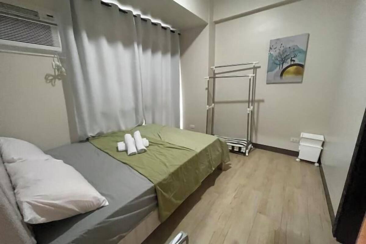 A single bed with a green bedspread, white pillows, and a white towel on the side. There is a window with curtains, a radiator, a wall-mounted towel rack, and a small white cabinet.