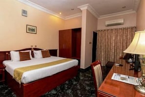 A hotel room with a large bed, a desk with a lamp, a chair, a wardrobe, and an air conditioning unit on the wall. - Sunfit International Limited (Lagos)