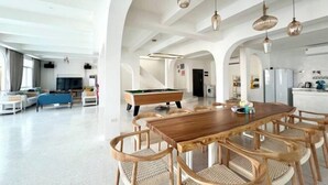 A spacious living area with a wooden dining table and chairs, a pool table, and a kitchen area visible through an archway. - Metero Garden Swimming Pool Villa (Pattaya)