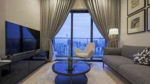 A modern living room with a large TV, a round coffee table with a plant, a sofa, and a chair, with a city view through large windows. - Lucentia Residence at Kuala Lumpur by Dreamcloud Sdn Bhd (Kuala Lumpur)