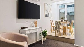 Una sala de estar moderna con un televisor de pantalla plana, un escritorio blanco, una silla cómoda, un comedor con sillas de madera y un ventanal grande.