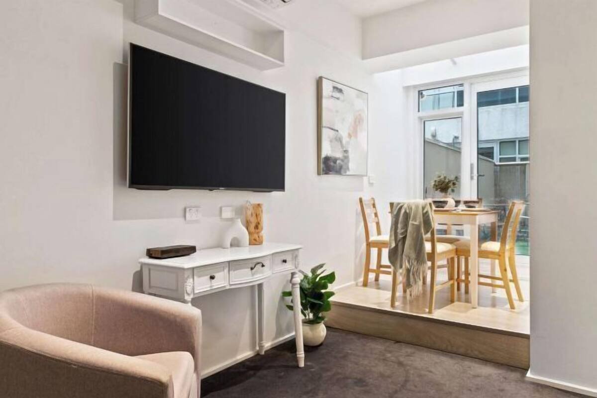 Una sala de estar moderna con un televisor de pantalla plana, un escritorio blanco, una silla cómoda, un comedor con sillas de madera y un ventanal grande.