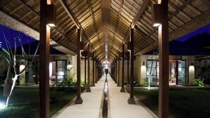 A covered walkway with a central water feature, flanked by columns and plants, leading to a building with large windows. - Villa Melissa (Bali)