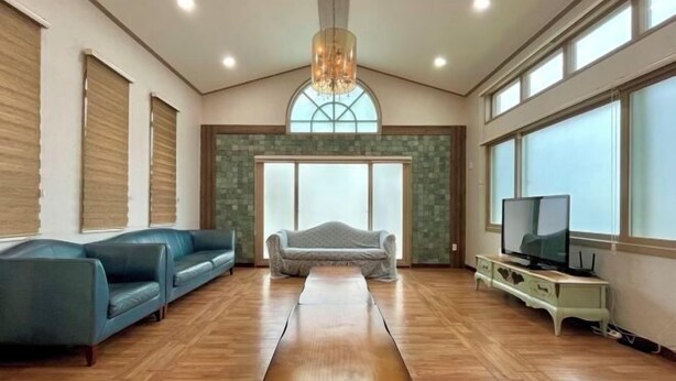 A spacious living room with a wooden floor, a large window, a chandelier, and a television. - Tebaz (Jeju)