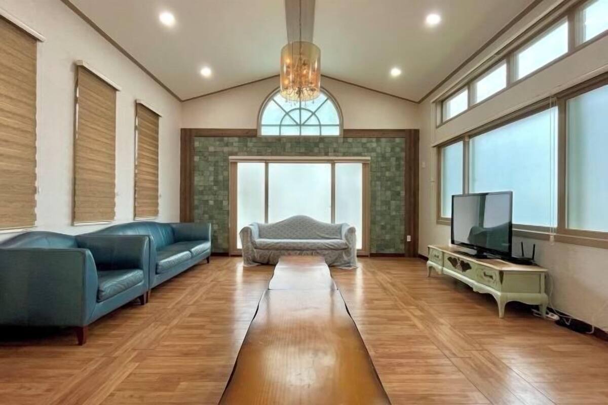 A spacious living room with a wooden floor, a large window, a chandelier, and a television.
