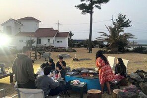 An outdoor dining area with a table set for a meal, surrounded by chairs and a few people. - Tebaz (Jeju)