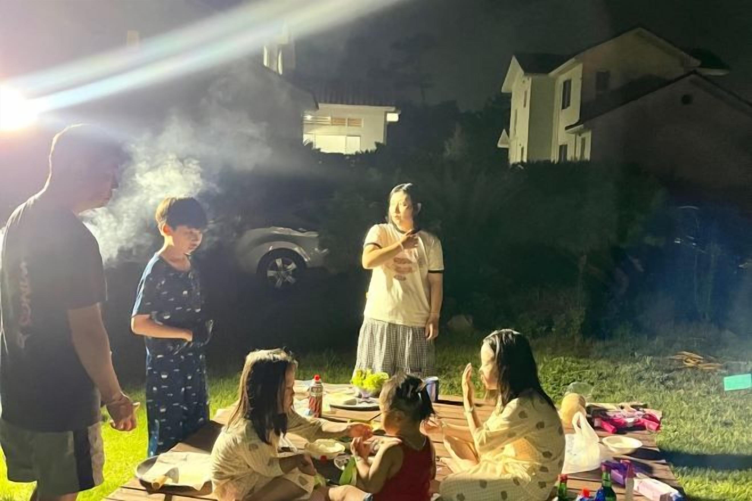 Un encuentro al aire libre con gente sentada en mesas, una persona de pie y una casa al fondo.