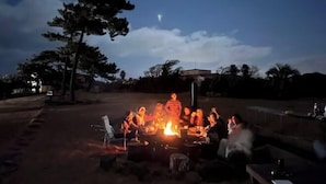 An outdoor gathering around a campfire with people seated on chairs, a table, and a few standing individuals. - Tebaz (Jeju)