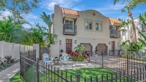 A two-story residential building with a tiled roof, a balcony, and a fenced backyard with a lawn and potted plants.