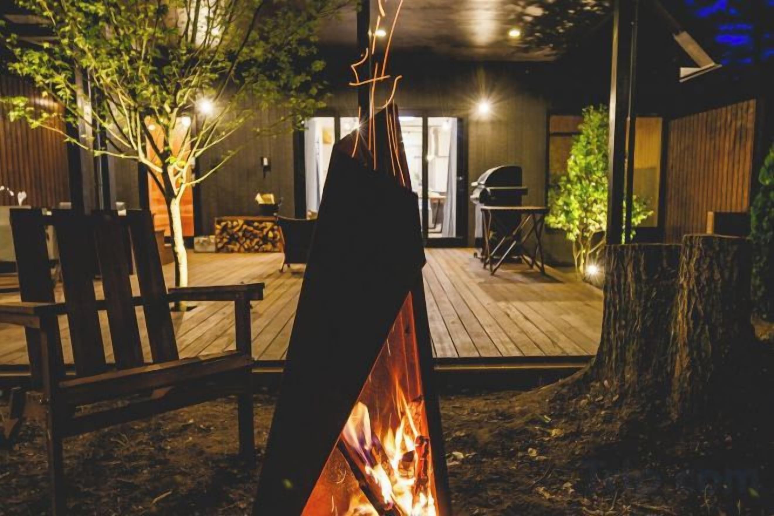 A fire pit in a backyard with a wooden deck, a barbecue grill, and a dining area with chairs.