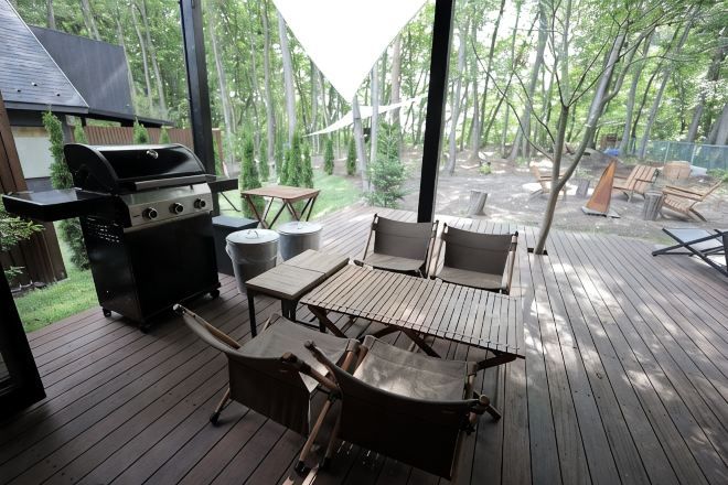 An outdoor deck with a grill, wooden table, and chairs, surrounded by trees.
