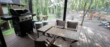 An outdoor deck with a grill, wooden table, and chairs, surrounded by trees.