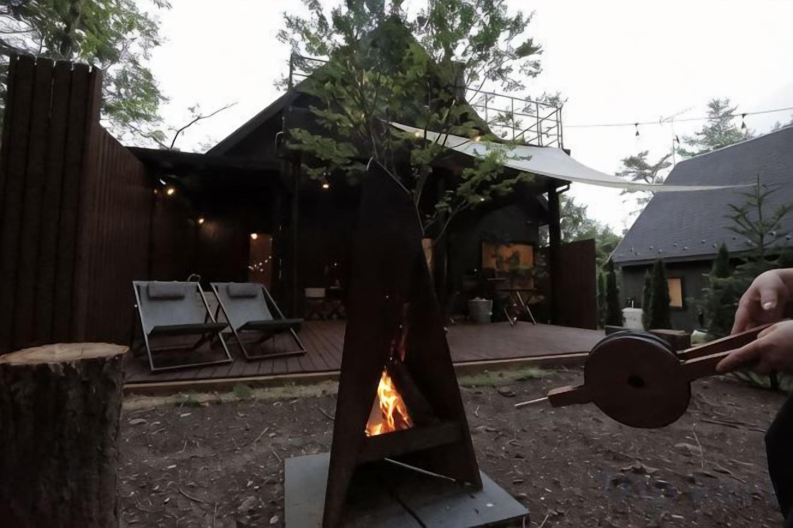 A metal fire pit with a wooden structure, surrounded by a wooden deck and a building with a sloped roof.