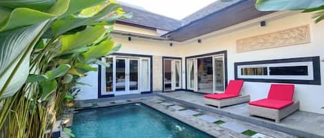 Una piscina con zona de patio, rodeada por una casa con puertas correderas de vidrio y sillas rojas acolchadas.