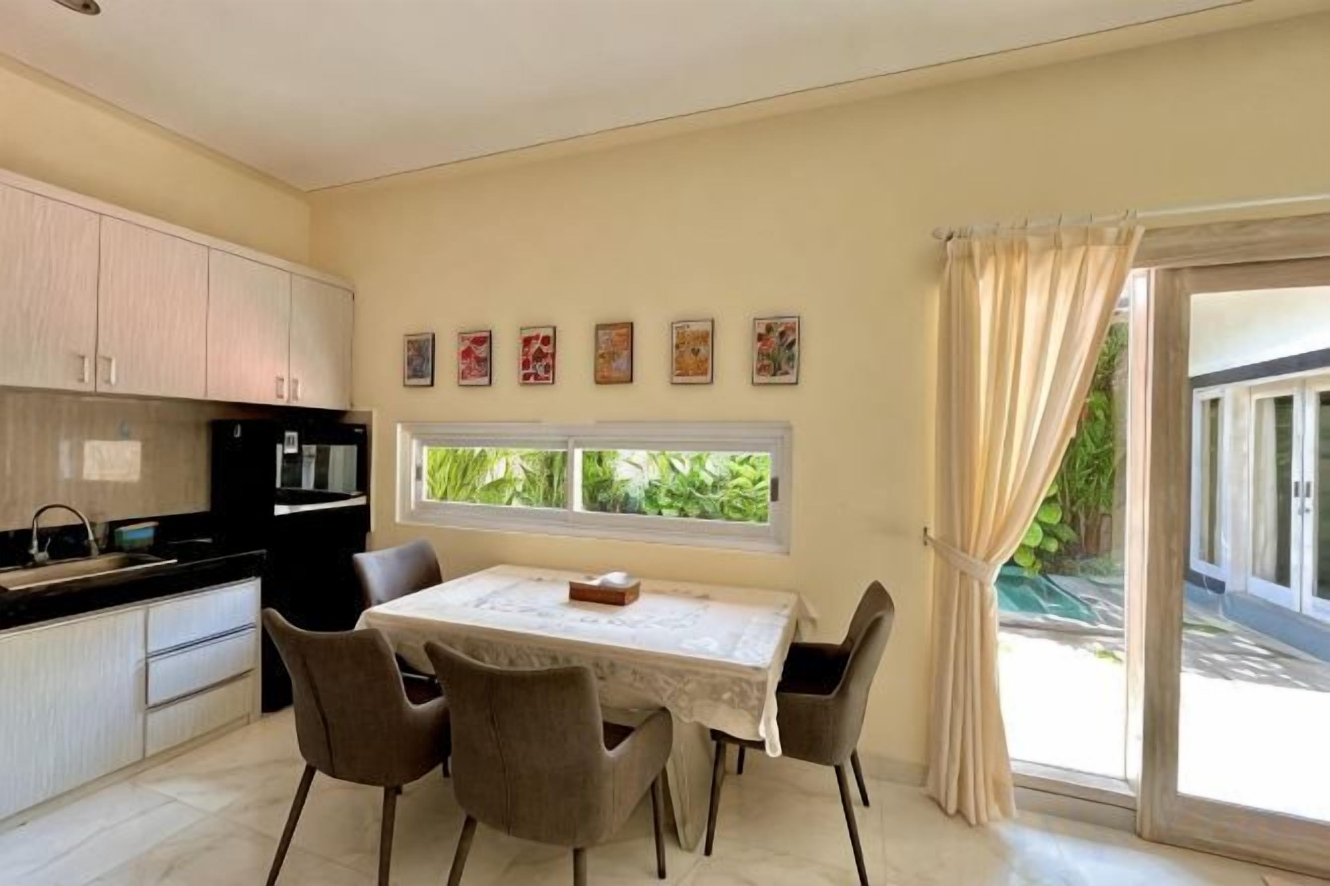 A dining area with a table and chairs, a kitchen with cabinets, a window with curtains, and a view of greenery outside.