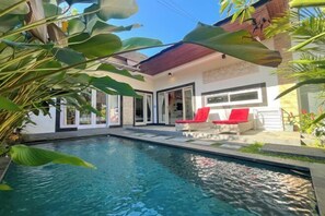A swimming pool with a patio area, a covered seating area, and a building with large windows and doors. - Enno Villa Sanur (Bali)