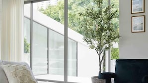 A modern living room with a white sofa, a dark armchair, and a large window offering a view of greenery outside. - Prime Beach House No. V7 (Sattahip)