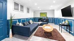 Un salon moderne avec un canapé d’angle, une table basse, un téléviseur à écran plat et, au fond, un coin cuisine.