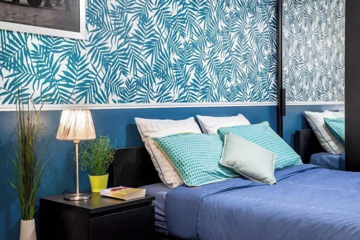 Een slaapkamer met een blauw sprei, meerdere kussens, een nachtkastje met een lamp en een muur met een bladmotief.