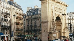 Une scène de rue avec une arche imposante, des bâtiments dotés de balcons et une enseigne indiquant « Café des Artistes ».