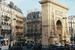 A street scene with a prominent archway, buildings with balconies, and a sign for 'Café des Artistes.' - Elegant 130m² Apartment in Central Paris, Marais (Paris)