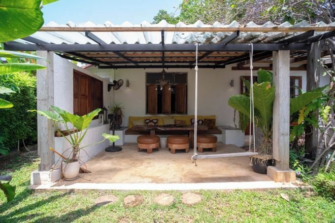 A covered outdoor seating area with a swing, surrounded by potted plants and a garden.