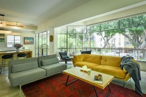A modern living room with a sofa, a coffee table, and a view of the outdoors through large windows. - Spacious & Green 3BR Apt w Balcony in City Center (Tel Aviv)