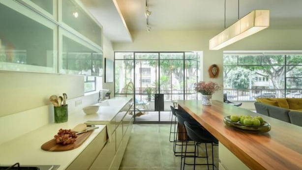 A modern kitchen with a wooden countertop, bar stools, and a large window offering a view of greenery outside. - Spacious & Green 3BR Apt w Balcony in City Center (Tel Aviv)