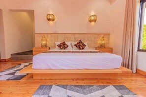 A bedroom with a large bed, two nightstands, decorative wall sconces, and a view of greenery outside.