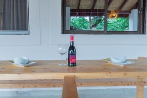 A wooden table with a bottle of wine and two glasses, set against a white wall with a window overlooking greenery. - Jandi Diani (Diani Beach)