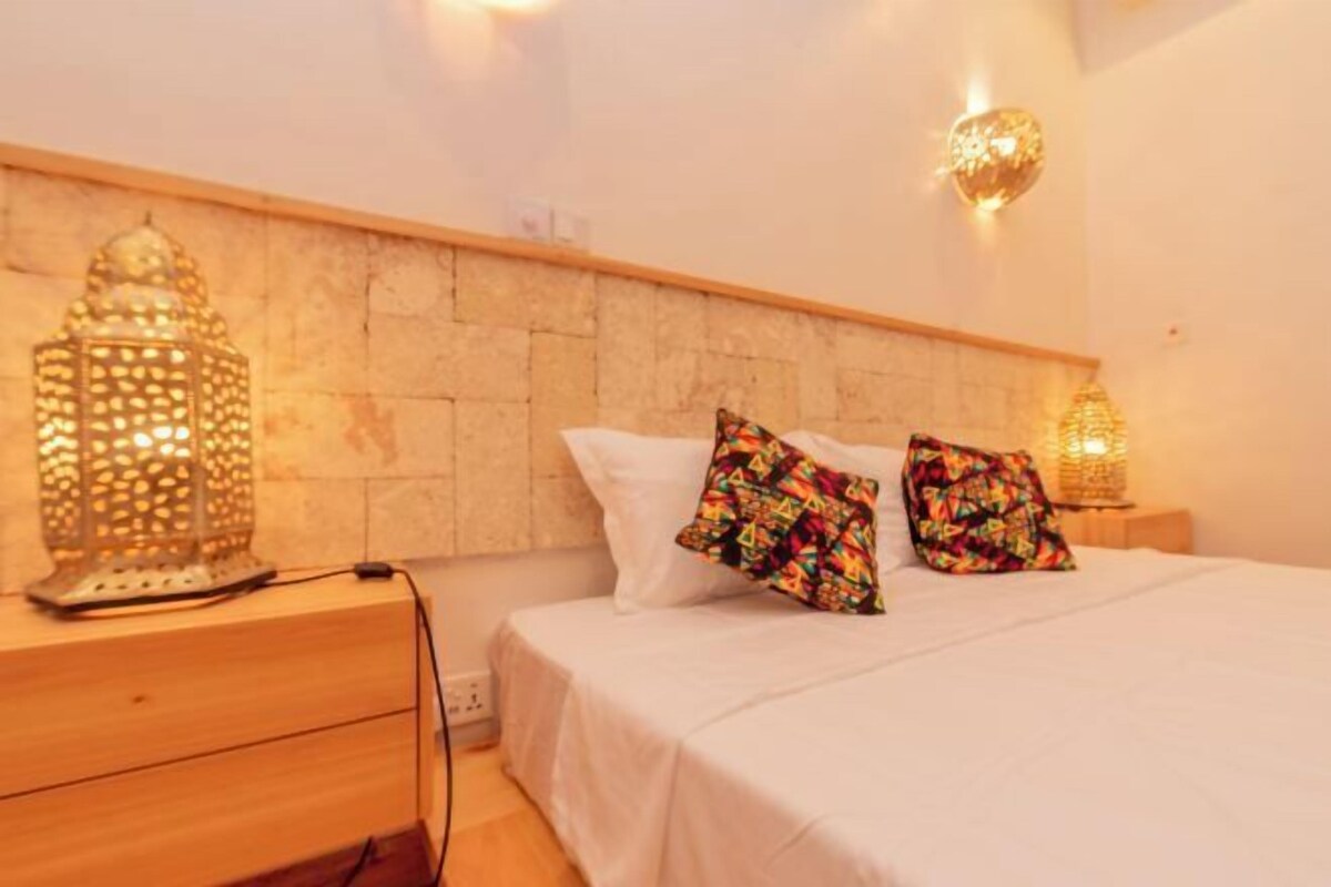 A neatly made bed with white bedding and colorful pillows, a wooden nightstand with a lamp, and a stone-patterned headboard.