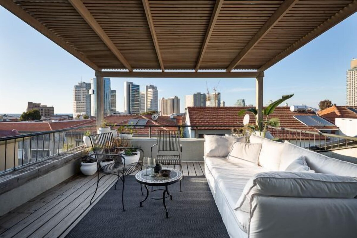 Una terraza en la azotea con una pérgola de madera, un sofá blanco, una mesa redonda y plantas en macetas.