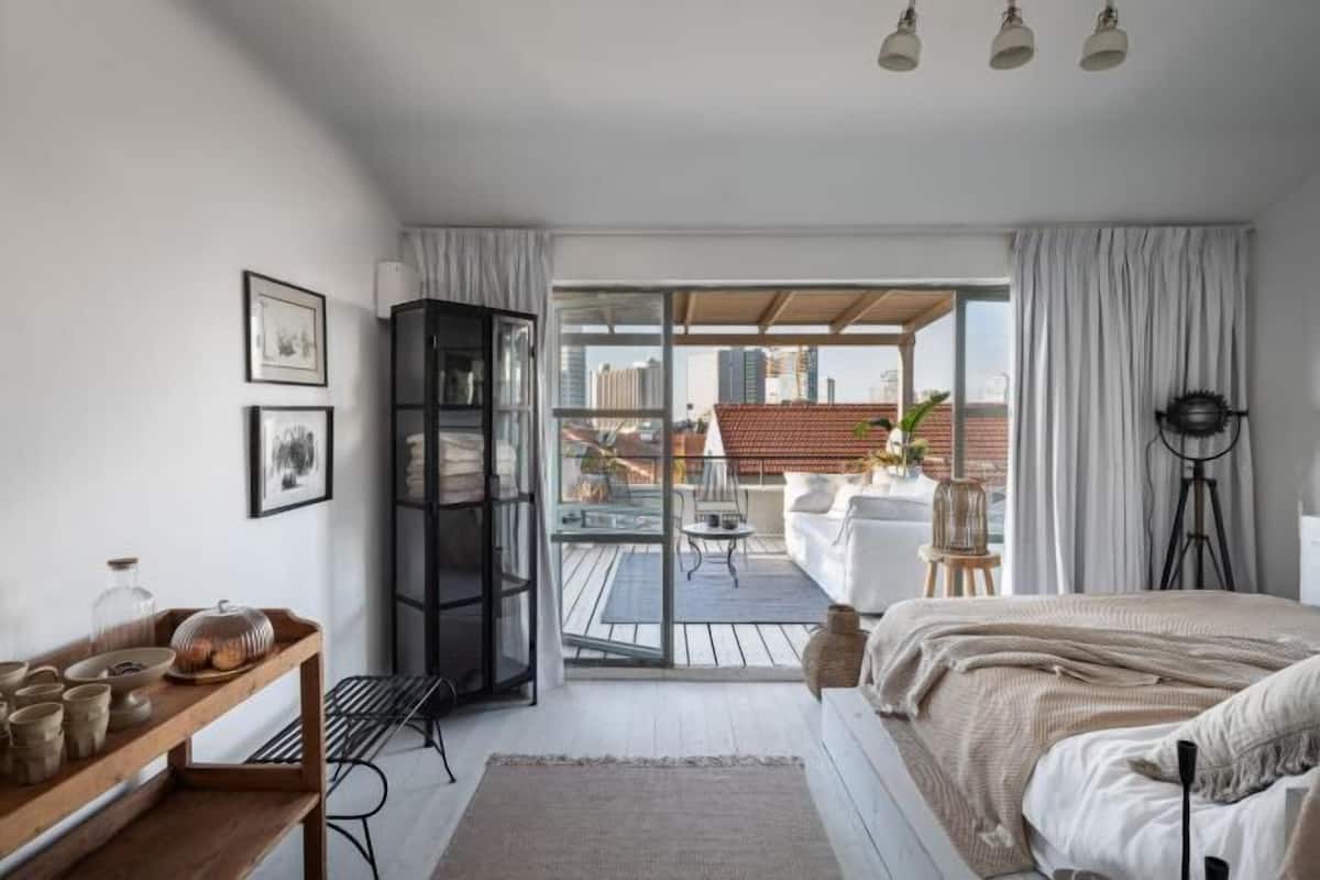Een slaapkamer met een bed, een houten nachtkastje, een zwarte plankkast, een glazen deur naar een balkon en uitzicht op de stad.