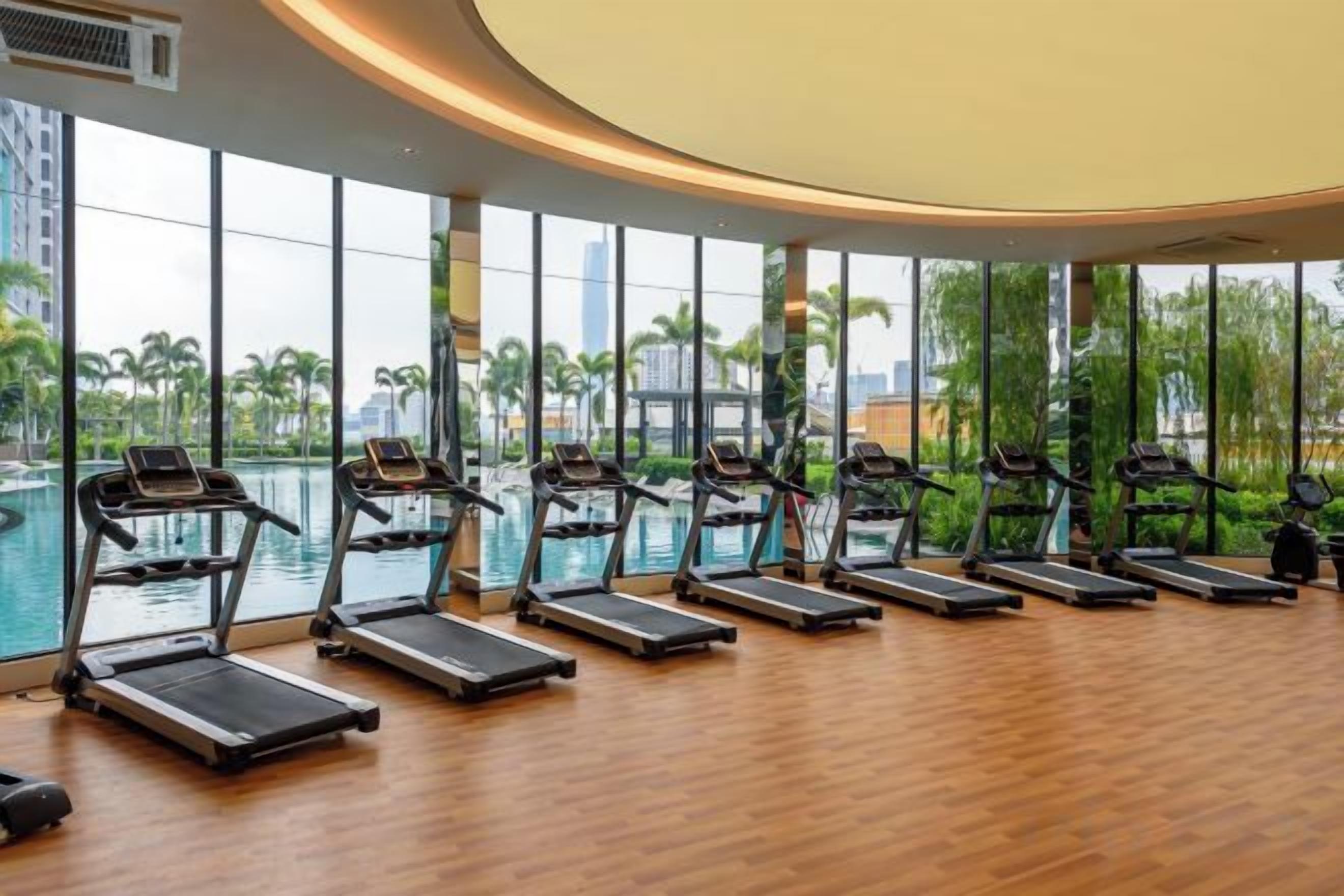 A gym with treadmills aligned against a wall with large windows overlooking a body of water and palm trees.
