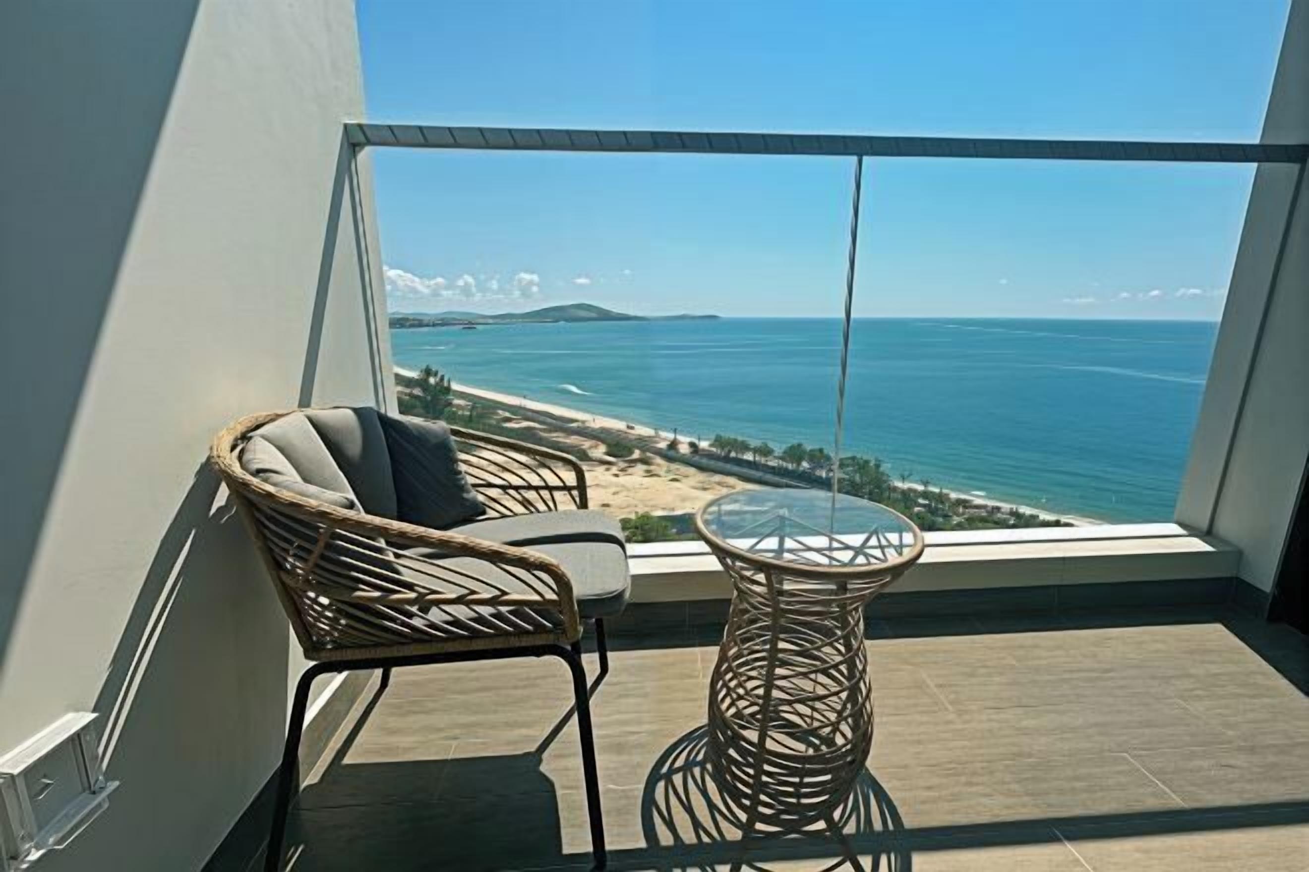Un balcón con mobiliario de mimbre, una mesa de vidrio y vista al océano y la playa.