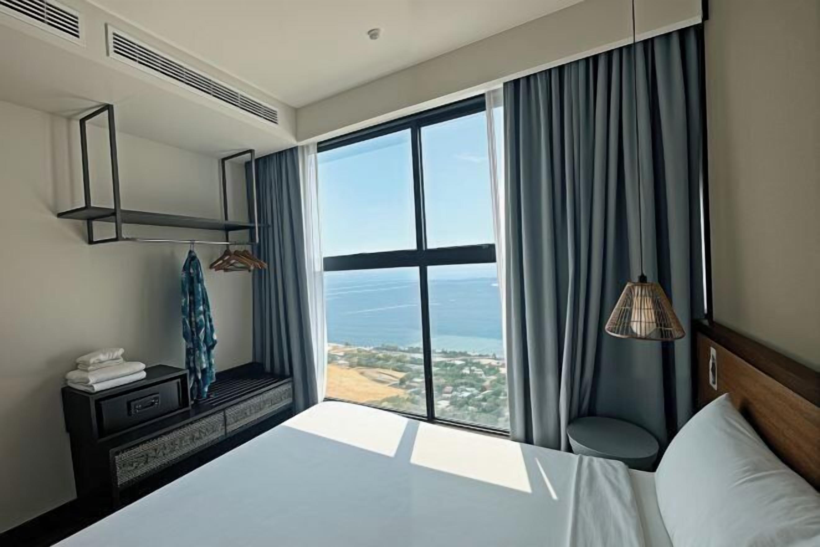 Habitación de hotel con un ventanal grande, una cama, una mesita de noche con lámpara y un espejo en la pared.