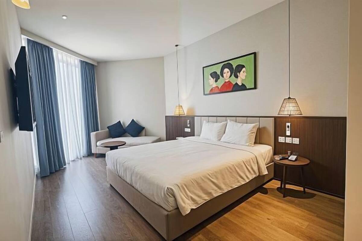 A hotel room with a bed, a sofa, a small table, a painting on the wall, and a window with curtains.