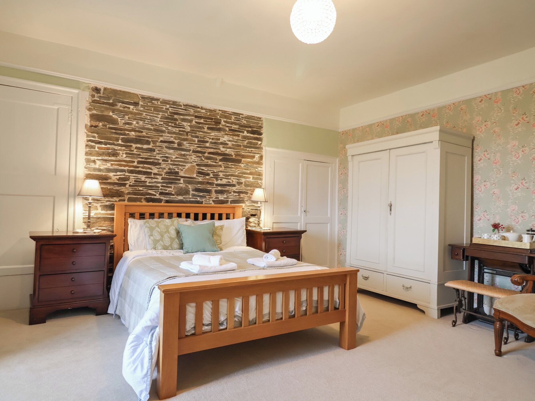 A bedroom with a stone wall, wooden bed, two bedside tables, a wardrobe, and a desk with a chair.