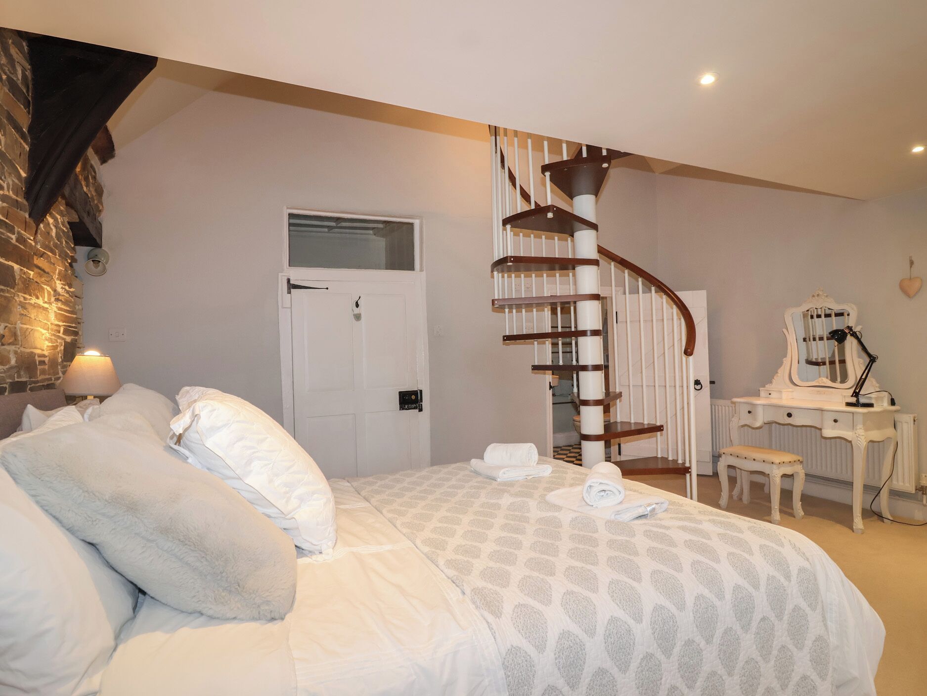 A bedroom with a bed, a white dresser with a mirror, a white chair, a spiral staircase, and a door.
