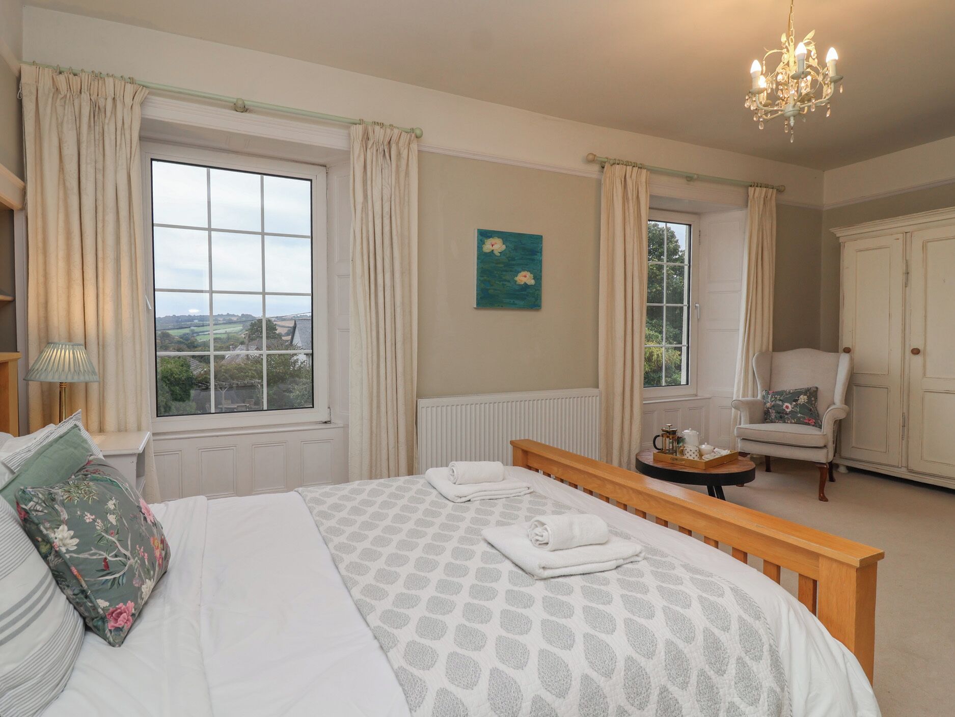 A bedroom with a large bed, two windows with curtains, a chandelier, a chair, and a cabinet.