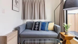 A modern living room with a grey sofa, patterned cushions, a glass coffee table, a side table with a plant, and a large window with curtains. - Higashi Mukoujima 6 chome (Tokyo)