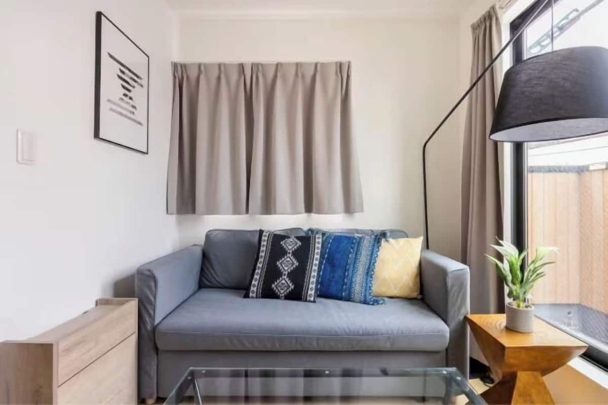 Un salon moderne avec un canapé gris, des coussins à motifs, une table basse en verre, une table d’appoint avec une plante et une grande fenêtre avec des rideaux.