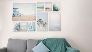 A wall with six framed beach scenes, a grey sofa with a green throw, and a single blue pillow. - The Garden Homestay BySTAY (Sekudai)