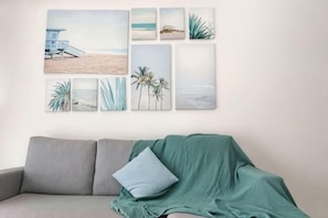 A wall with six framed beach scenes, a grey sofa with a green throw, and a single blue pillow. - The Garden Homestay BySTAY (Sekudai)