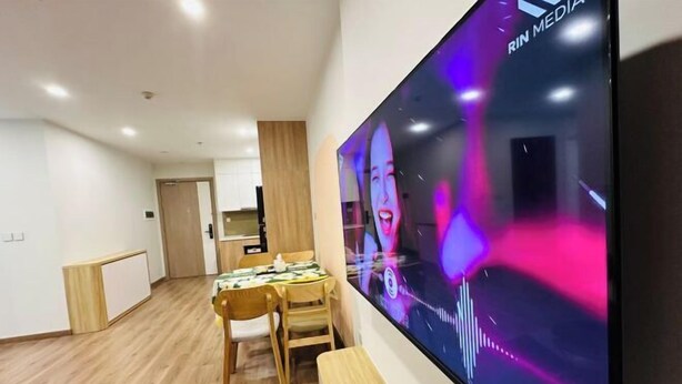 A modern living room with a large flat-screen TV mounted on the wall, a wooden dining table with chairs, and a kitchen area in the background. - 2BRS@Vinhomes Smart City (Hanoi)